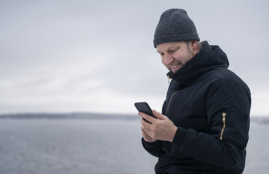 Mann med lue ser på mobilen sin ved vannet.